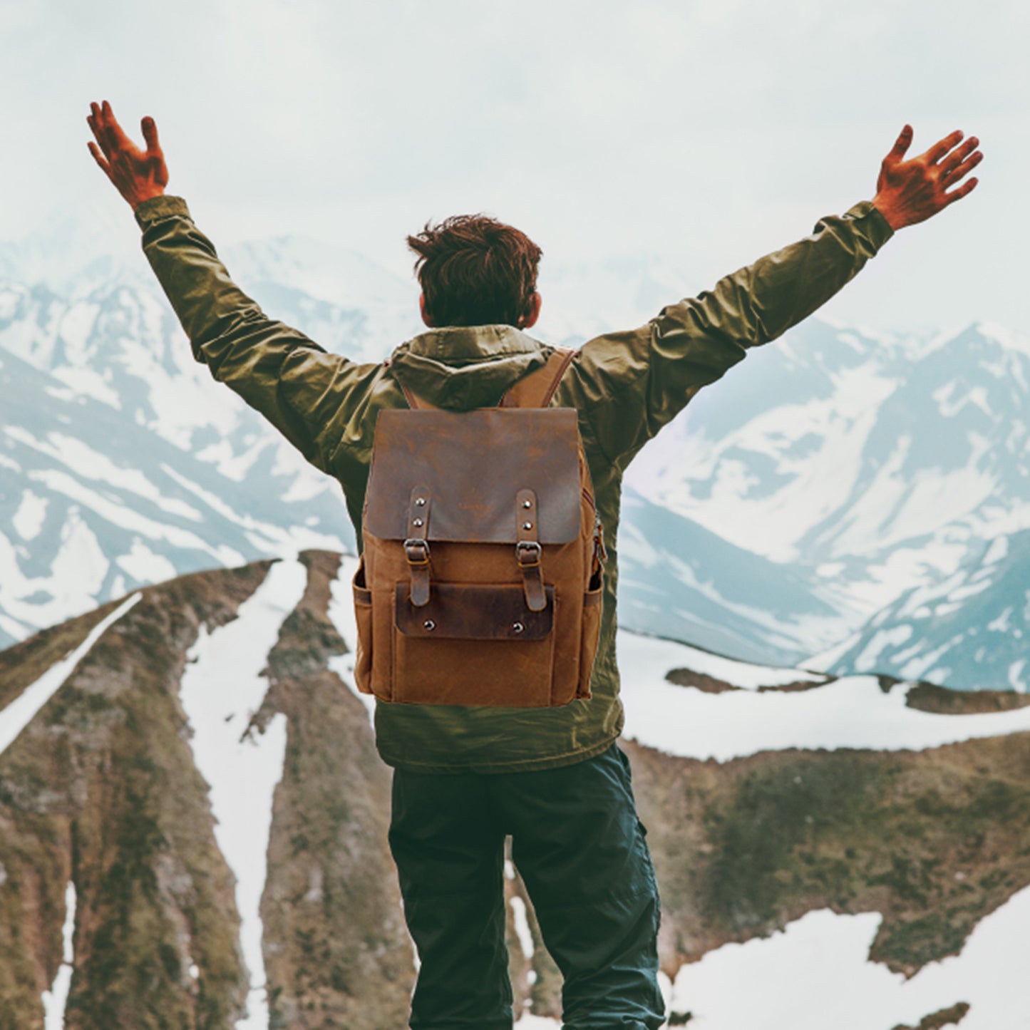 Person with a brown backpack standing with arms outstretched against a mountainous landscape