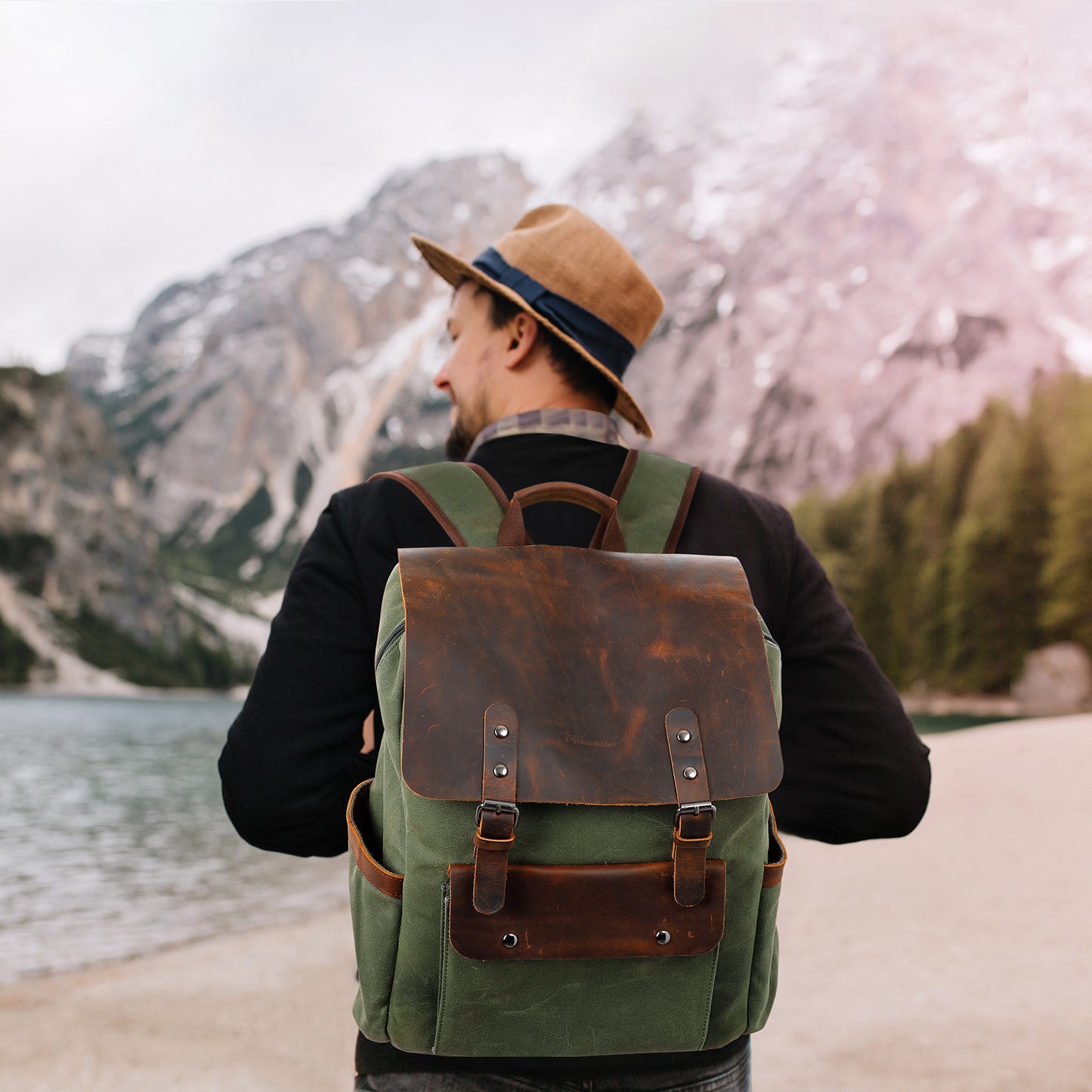 Man with a green backpack and brown hat standing by a lake with mountains in the background