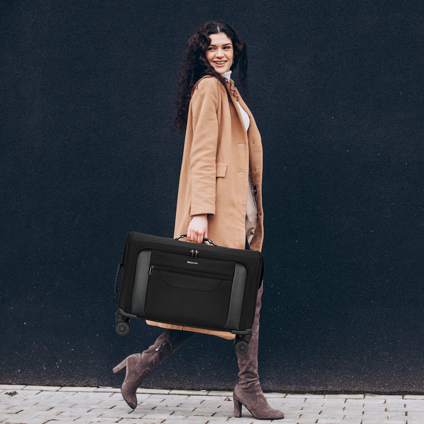 Woman in a tan coat holding a black suitcase against a dark background